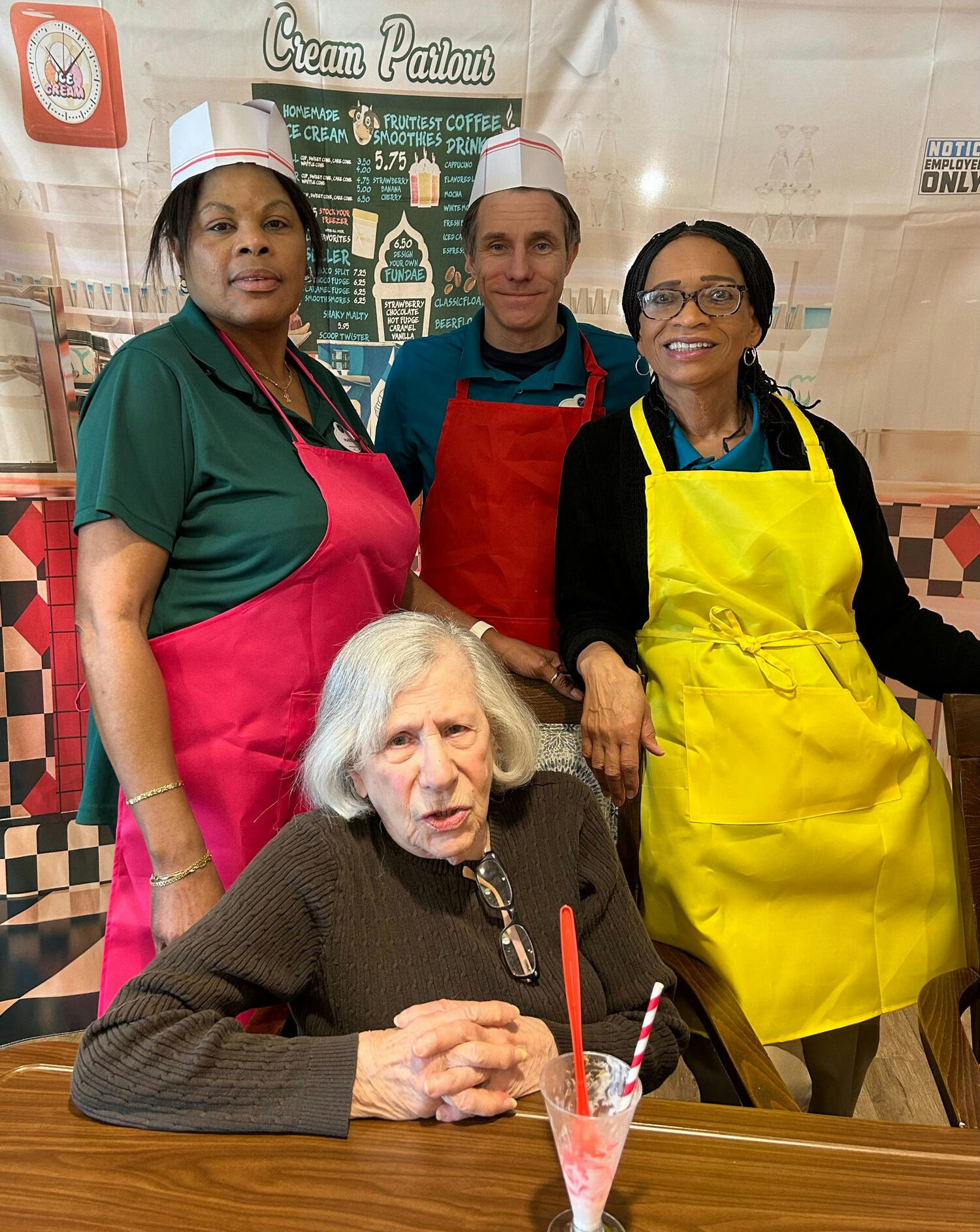 Three smiling staff members in colorful aprons gather around a seated resident enjoying a cold treat at an ice-cream-parlor-style spot inside Arbor Terrace Norwood, a senior living community in Norwood, NJ.