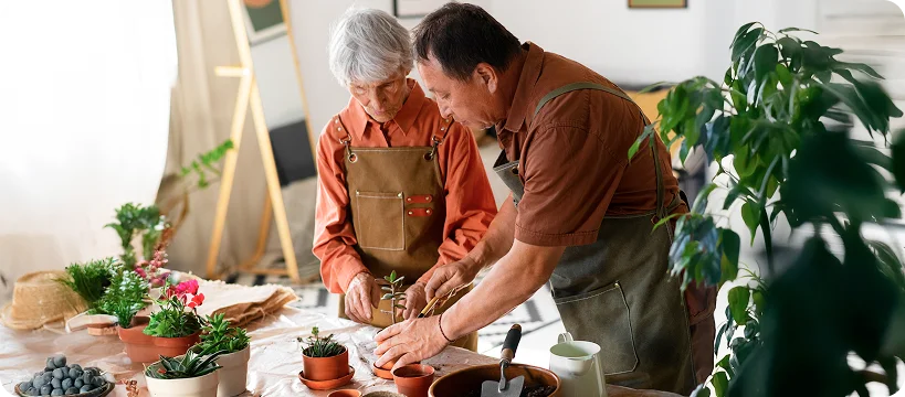 A senior couple in aprons works together on a potted plant at a table covered in gardening supplies.
